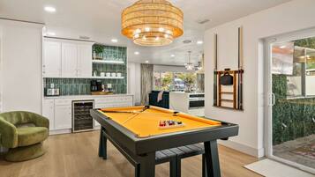 A modern indoor pool table with a pool cue, surrounded by a kitchen area with white cabinets and a green backsplash.