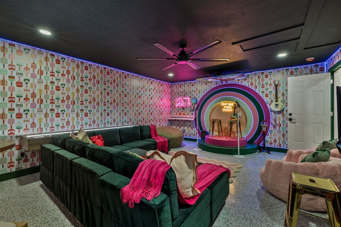 Une pièce avec un canapé vert, un fauteuil rose, un panneau lumineux au néon et un mur à motifs.