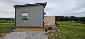A small, single-story cabin with a window and a door, situated on a gravel path with a grassy field in the background. - Whitetail Suites Peaceful Cabin 1, Near Amish Country & Mohican (Loudonville)