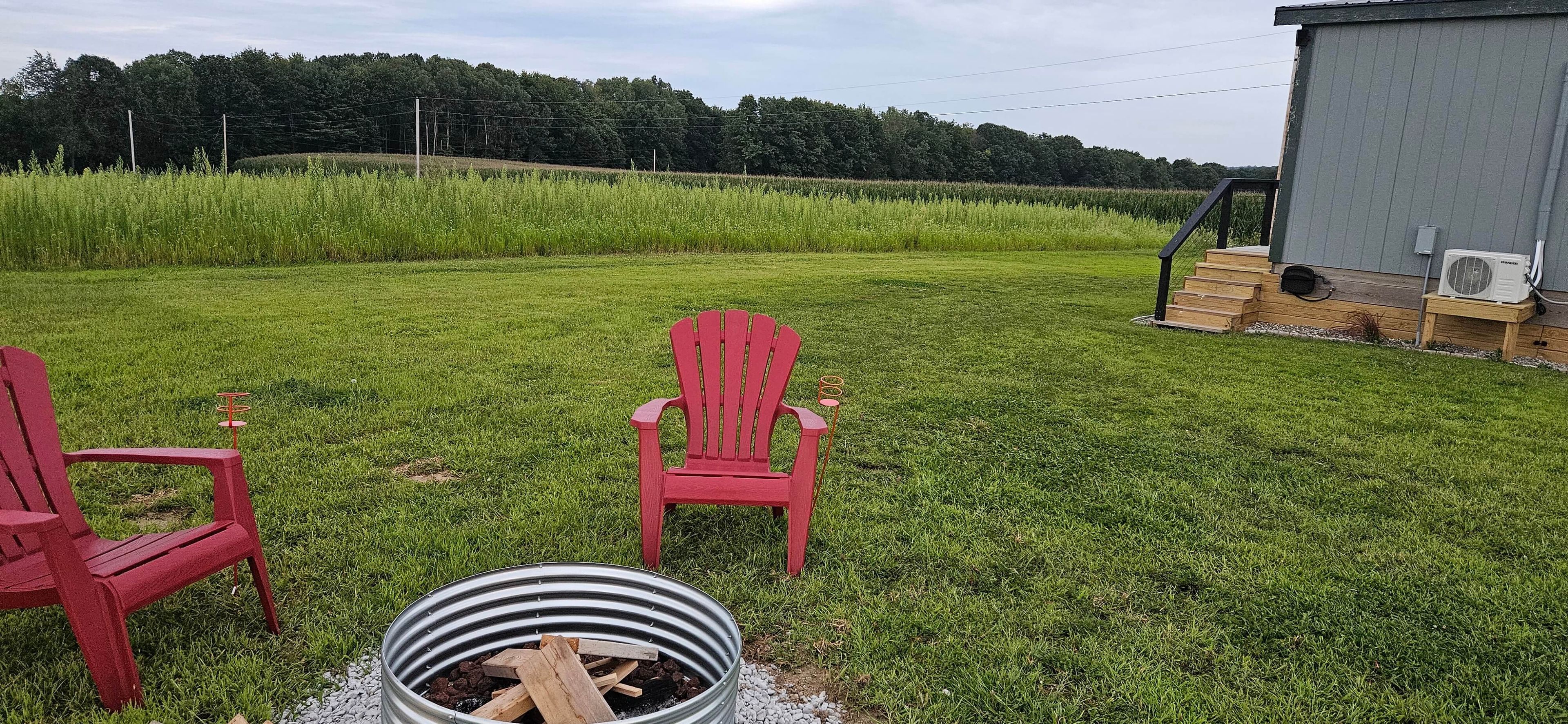 A grassy outdoor area with two red Adirondack chairs, a metal fire pit, and a wooden deck with a step leading to a building.