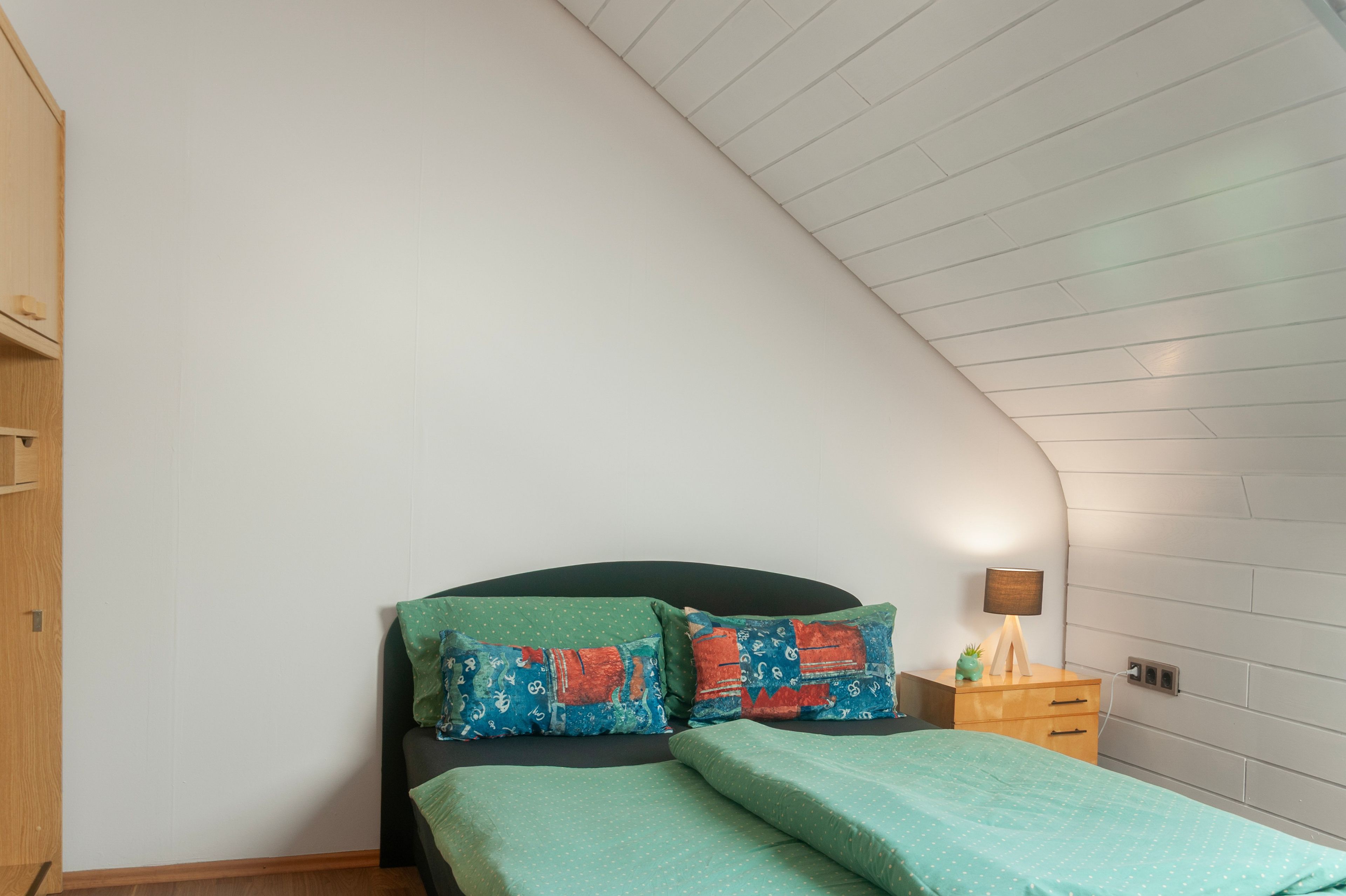 A bedroom with a sloped ceiling, a bed with green bedding, two pillows with colorful patterns, a bedside table with a lamp, and a wooden wardrobe.