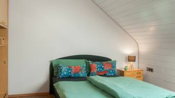 A bedroom with a sloped ceiling, a bed with green bedding, two pillows with colorful patterns, a bedside table with a lamp, and a wooden wardrobe.