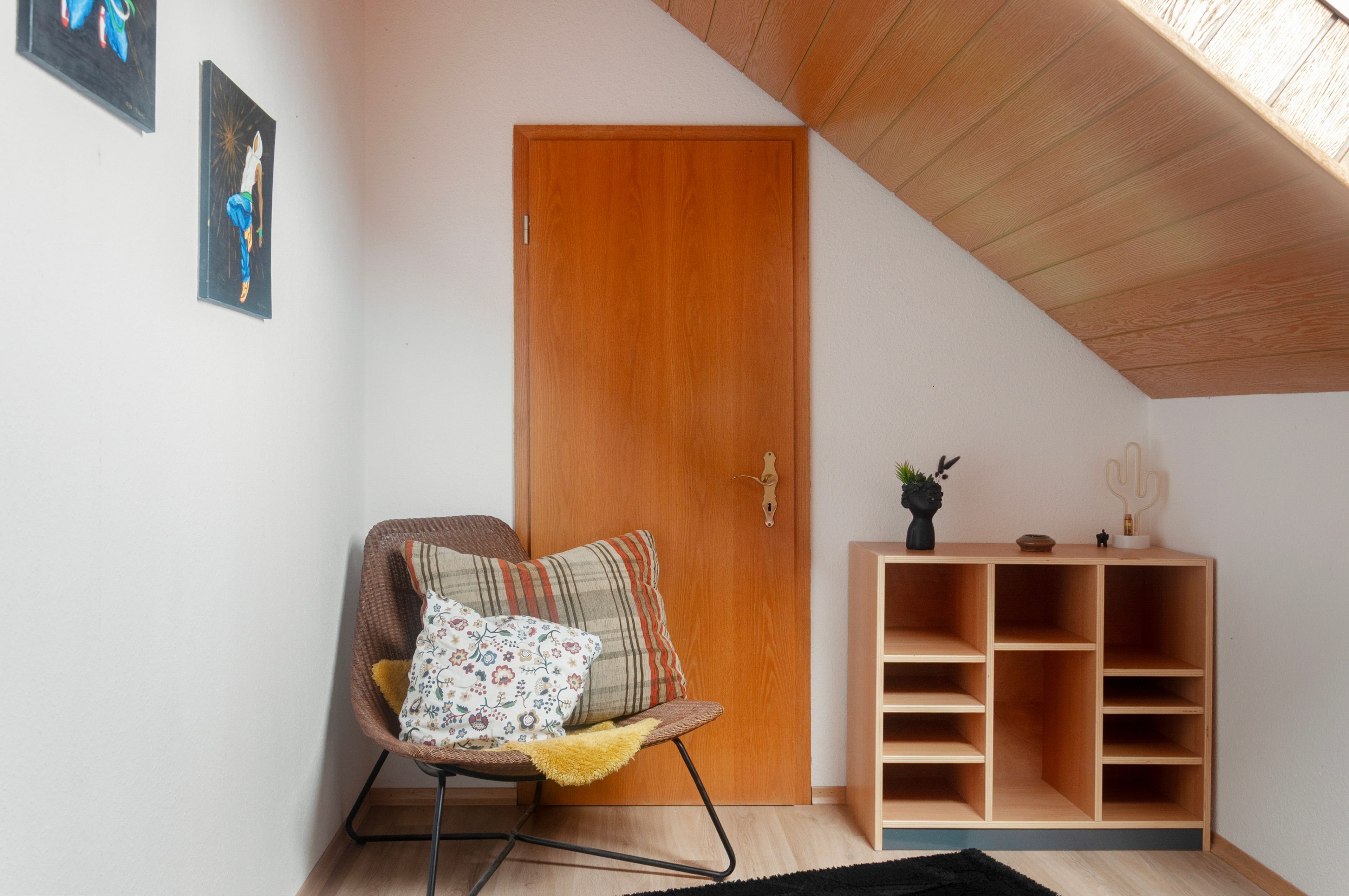 Une pièce avec une porte en bois, une chaise en osier dotée d’un coussin, une petite étagère en bois ornée d’objets décoratifs et un tapis noir sur le sol.