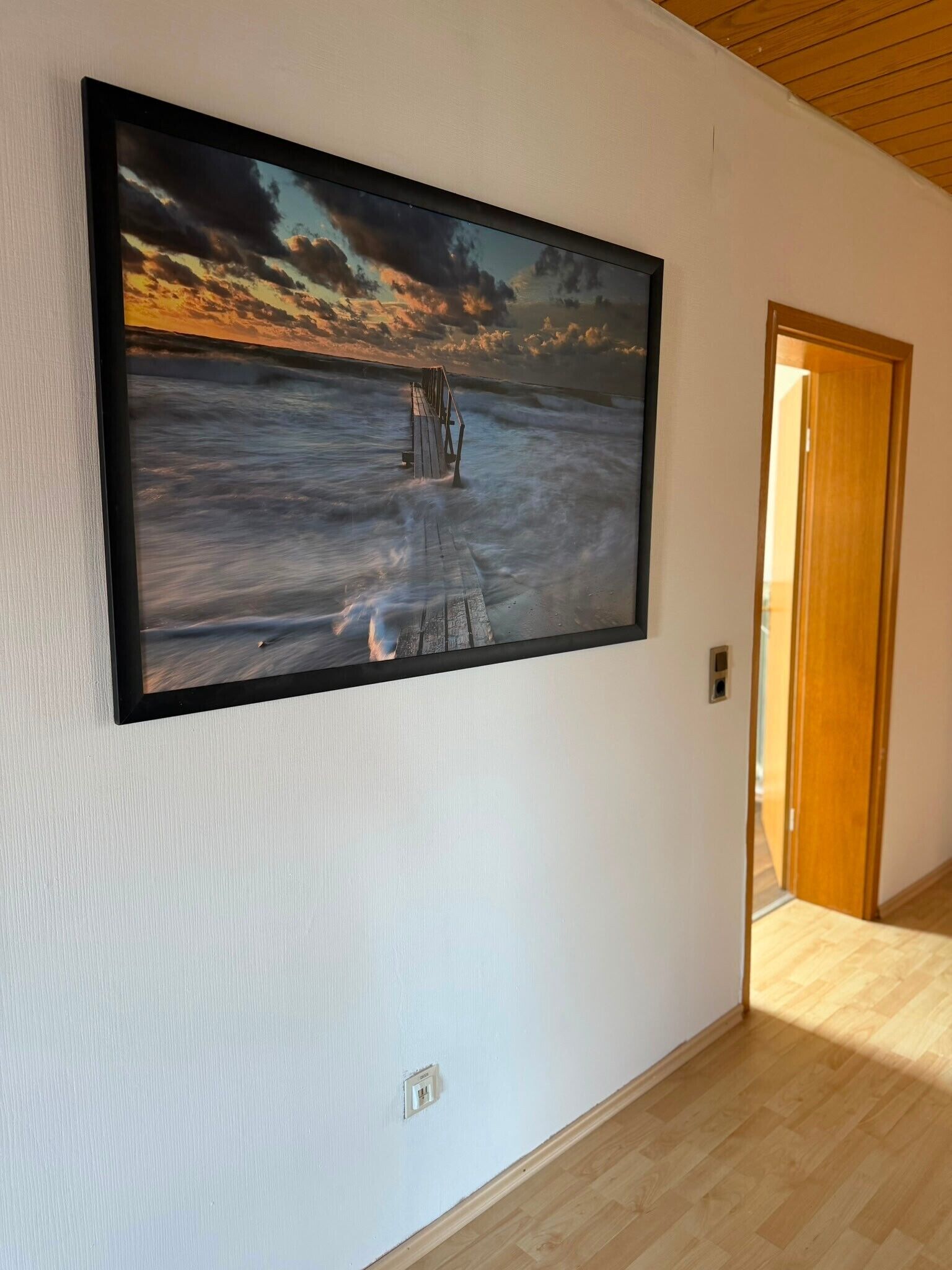 A hallway with a large framed artwork depicting a sunset over the ocean, a wooden door, and a light switch on the wall.