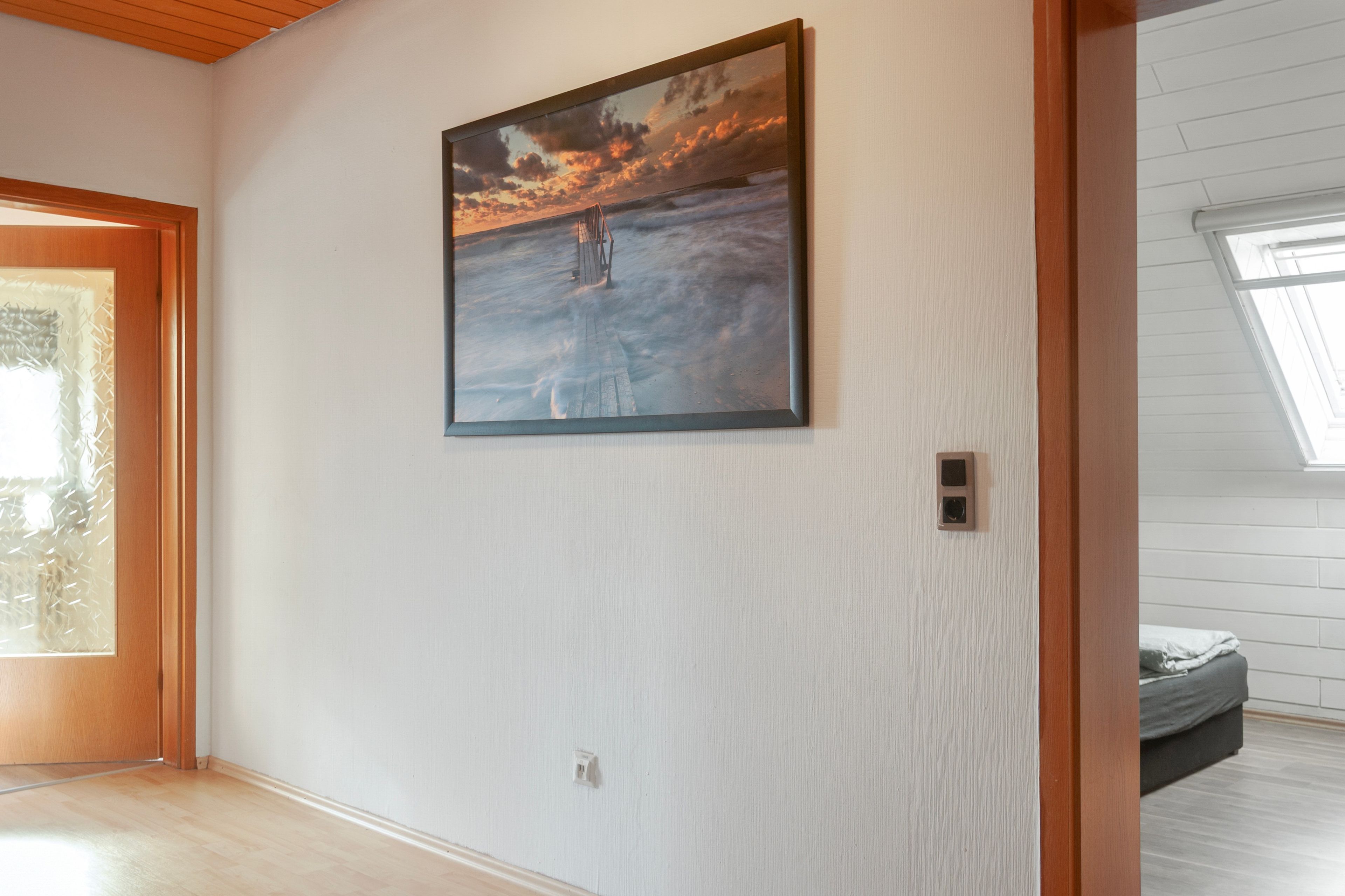A room with a painting of a sunset over the ocean, a wooden door, and a window with a view.