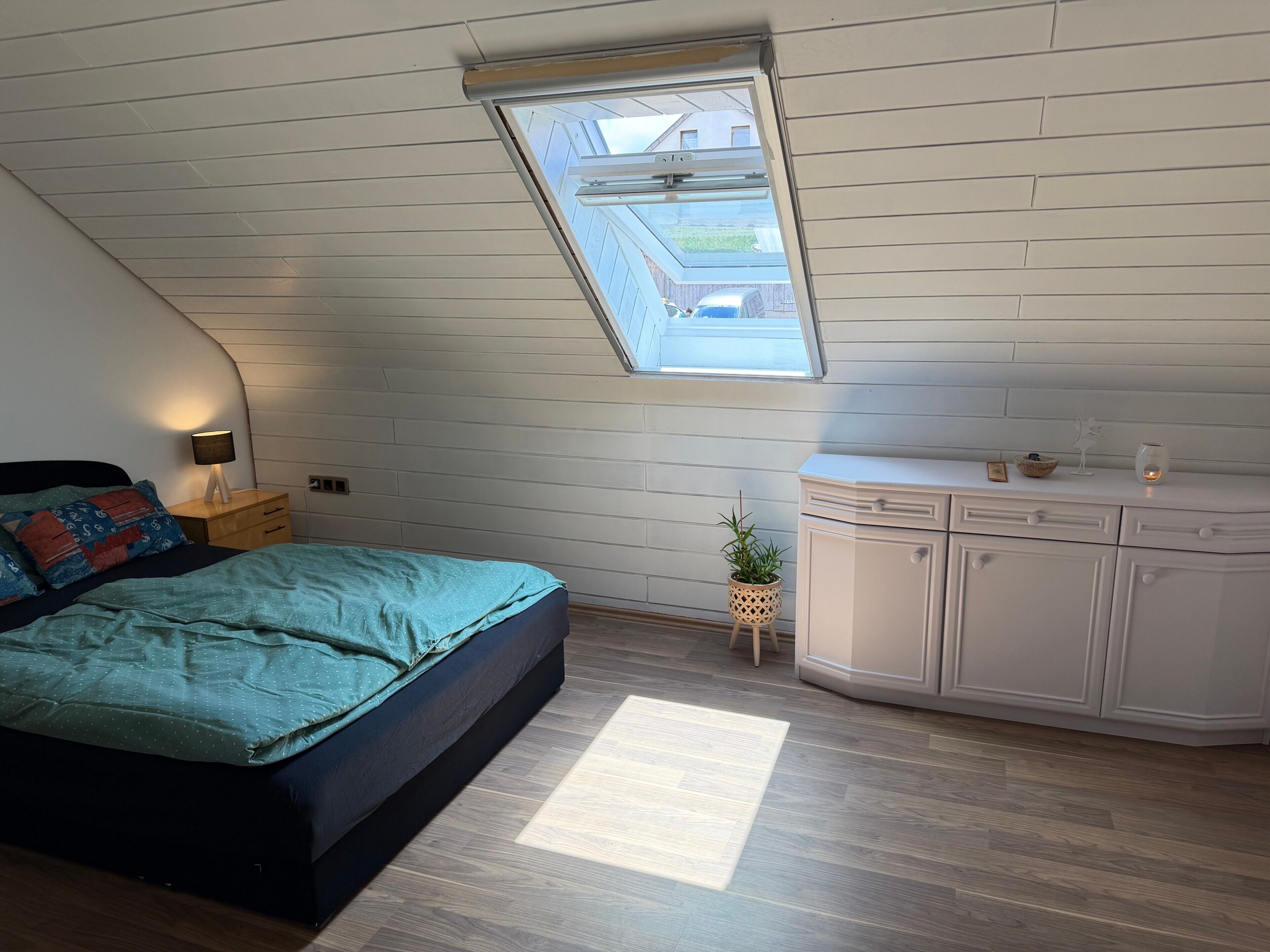 Quarto com teto inclinado, cama com colcha azul-turquesa, mesa de cabeceira com abajur, uma pequena planta em vaso e cómoda branca com gavetas.