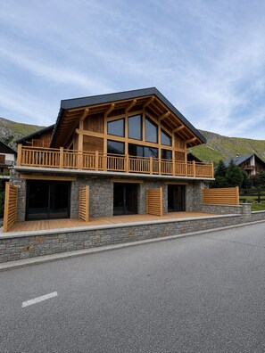 A two-story wooden cabin with a balcony, stone foundation, and a mountain backdrop. - At Ginou - Jogidi Chalet (Saint-Sorlin-d'Arves)