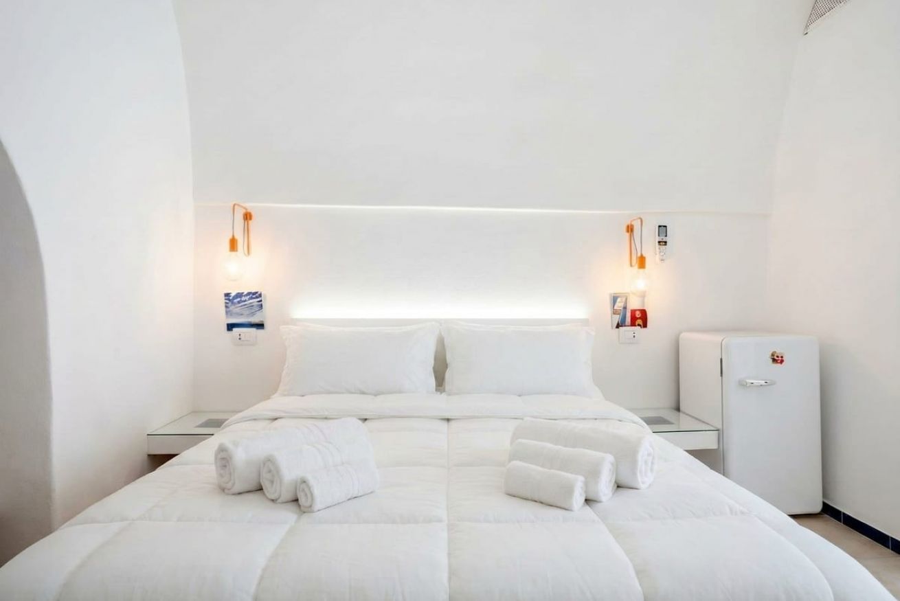 A modern hotel room with a large bed, two bedside tables, a small refrigerator, and wall-mounted lighting.