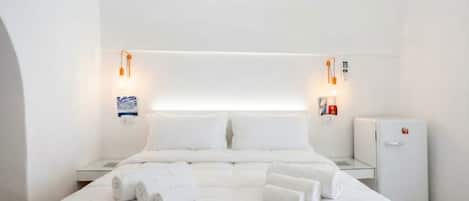 A modern hotel room with a large bed, two bedside tables, a small refrigerator, and wall-mounted lighting.