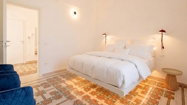 Quarto com cama, uma cadeira azul, uma mesinha redonda e piso estampado.