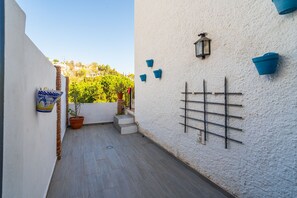 Un estrecho pasillo exterior con una pared blanca texturizada, una planta en maceta y un estante metálico.