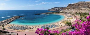 Uma praia costeira com uma linha de costa curva, um calçadão com guarda-sóis e um penhasco com edifícios.