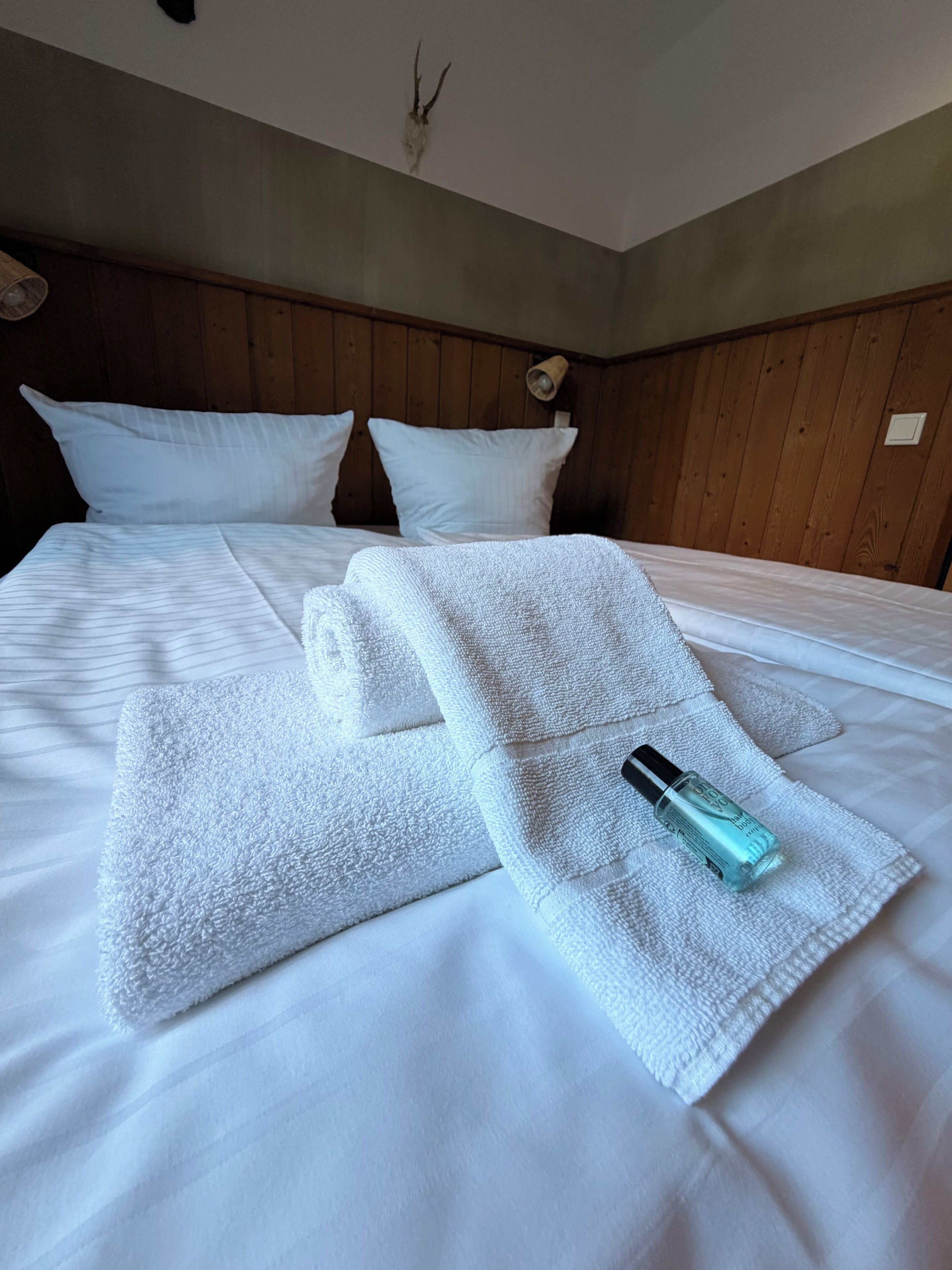 Ein ordentlich bezogenes Bett mit weißen Leinen und einem gefalteten Handtuch, eine Lotionflasche und eine Wandleuchte.