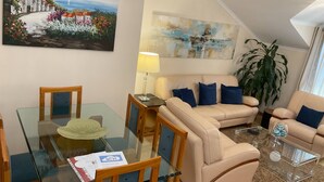 Sala de estar com mesa de jantar de vidro, sofá bege e uma pintura de uma paisagem costeira.