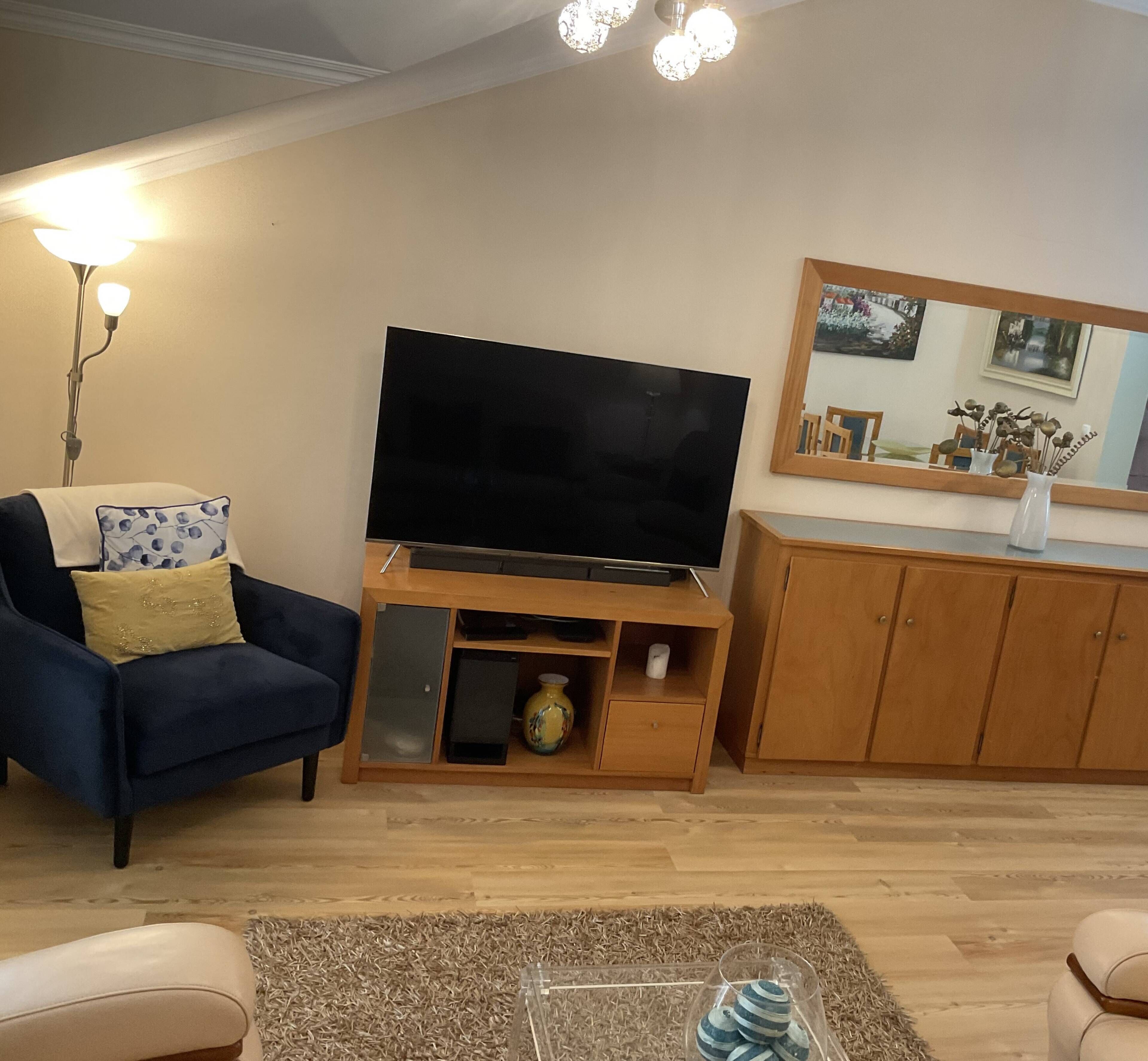 A living room with a flat-screen TV, a blue armchair, a wooden cabinet, and a mirror on the wall.