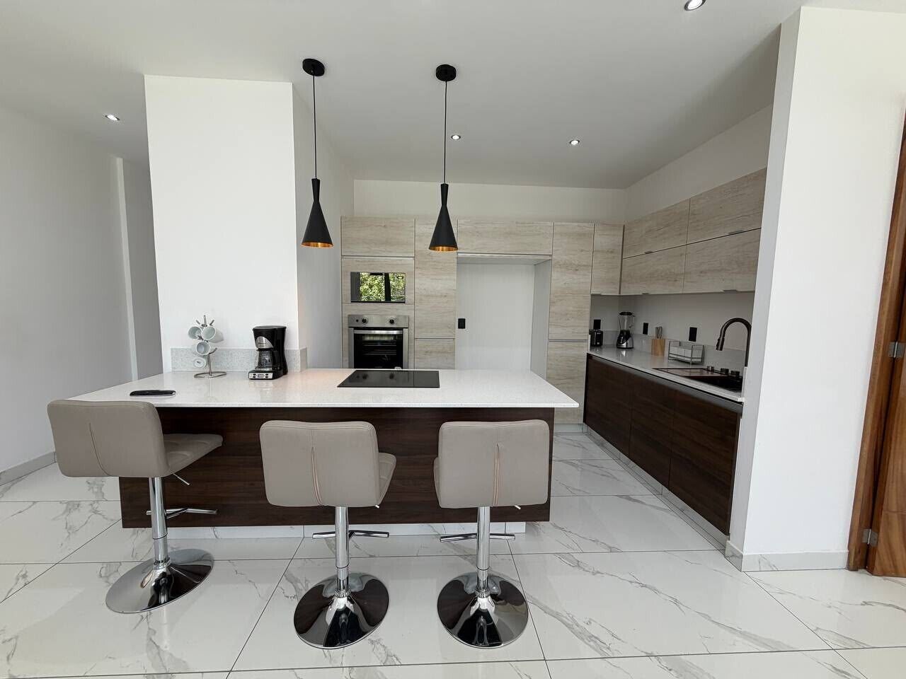 Una cocina moderna con una isla central, taburetes de bar, iluminación colgante y un horno empotrado.