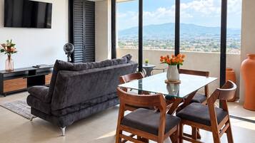 Un salon moderne comprenant un canapé, une table à manger en verre avec des chaises, un téléviseur à écran plat fixé au mur et une grande fenêtre offrant une vue sur la ville.