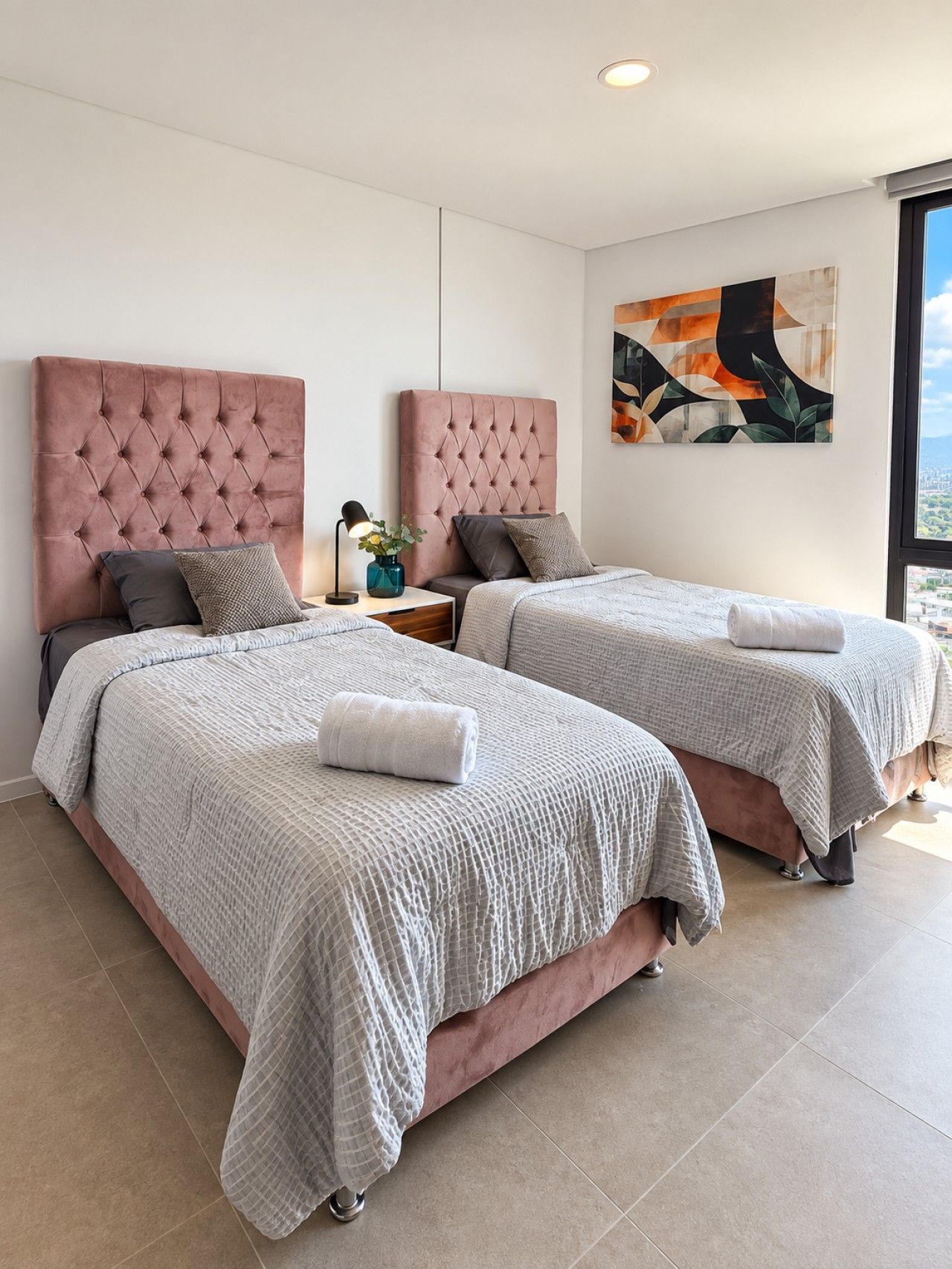 A modern bedroom with two beds, a nightstand, a lamp, and a painting on the wall.