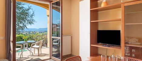 A room with a TV on a wooden shelf, a dining table with chairs, and a view of the outdoors through a glass door.