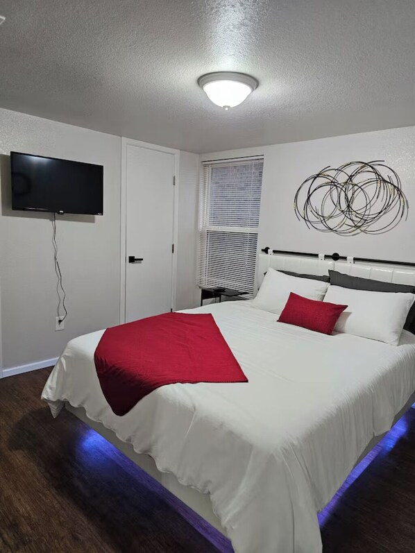 A bedroom with a bed, a television mounted on the wall, a window with blinds, and a ceiling light.
