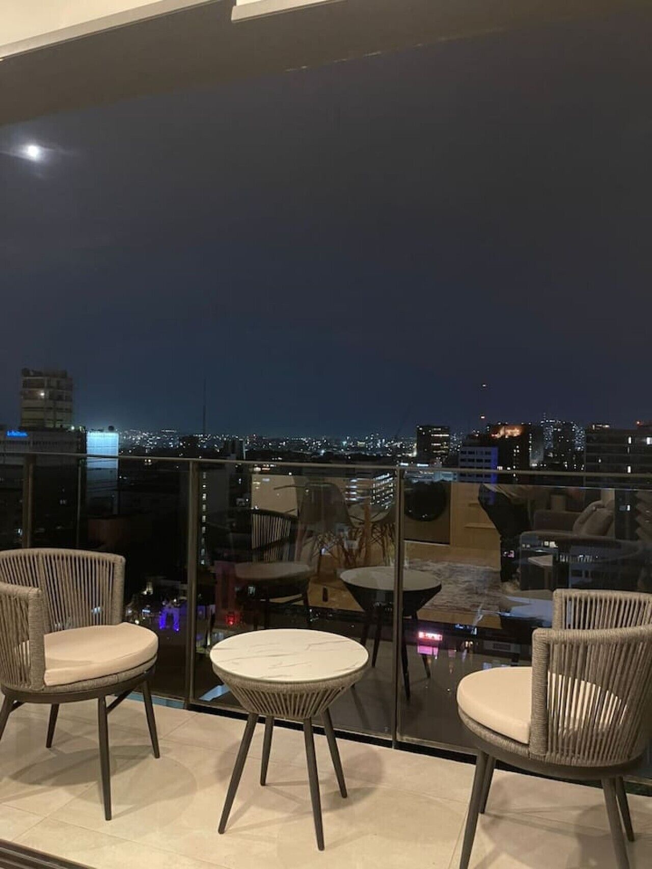 Un balcón con sillas de mimbre y una mesa de vidrio, con vista a un paisaje urbano nocturno.