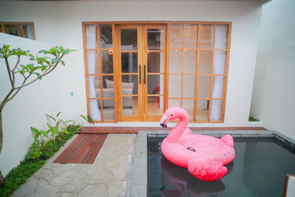 Een roze opblaasflamingo in een zwembad, met een houten deur en schuiframen op de achtergrond.