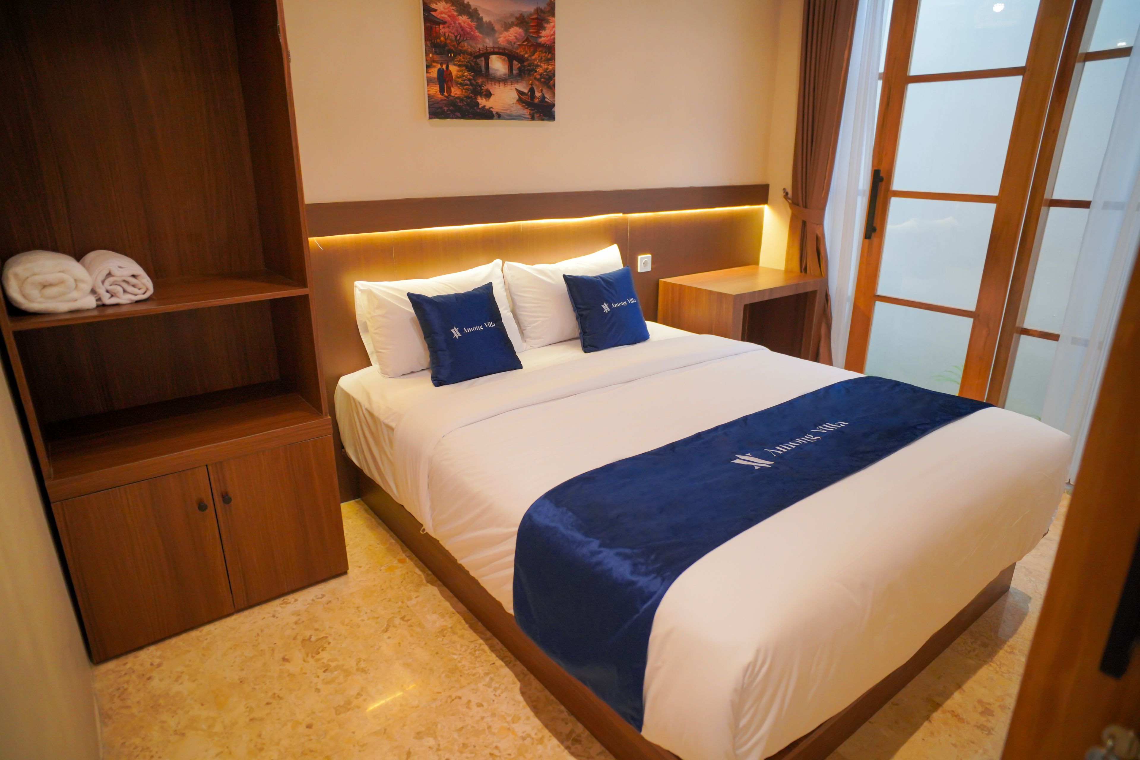 A hotel room with a bed, a headboard with built-in lighting, a nightstand, a wooden wardrobe, and a painting on the wall.