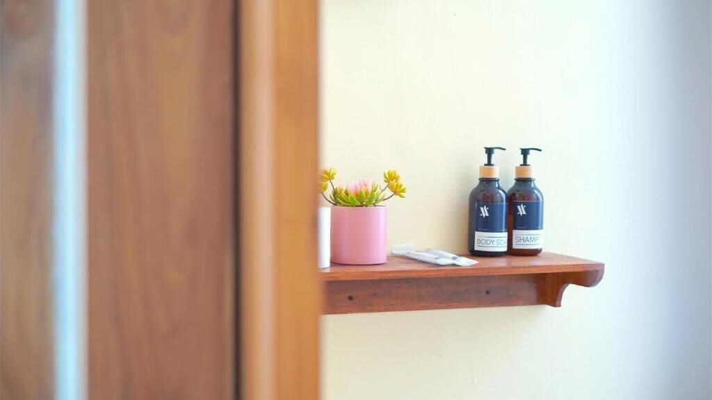 Una mensola in legno con un vaso rosa contenente fiori e due flaconi di spray corpo.