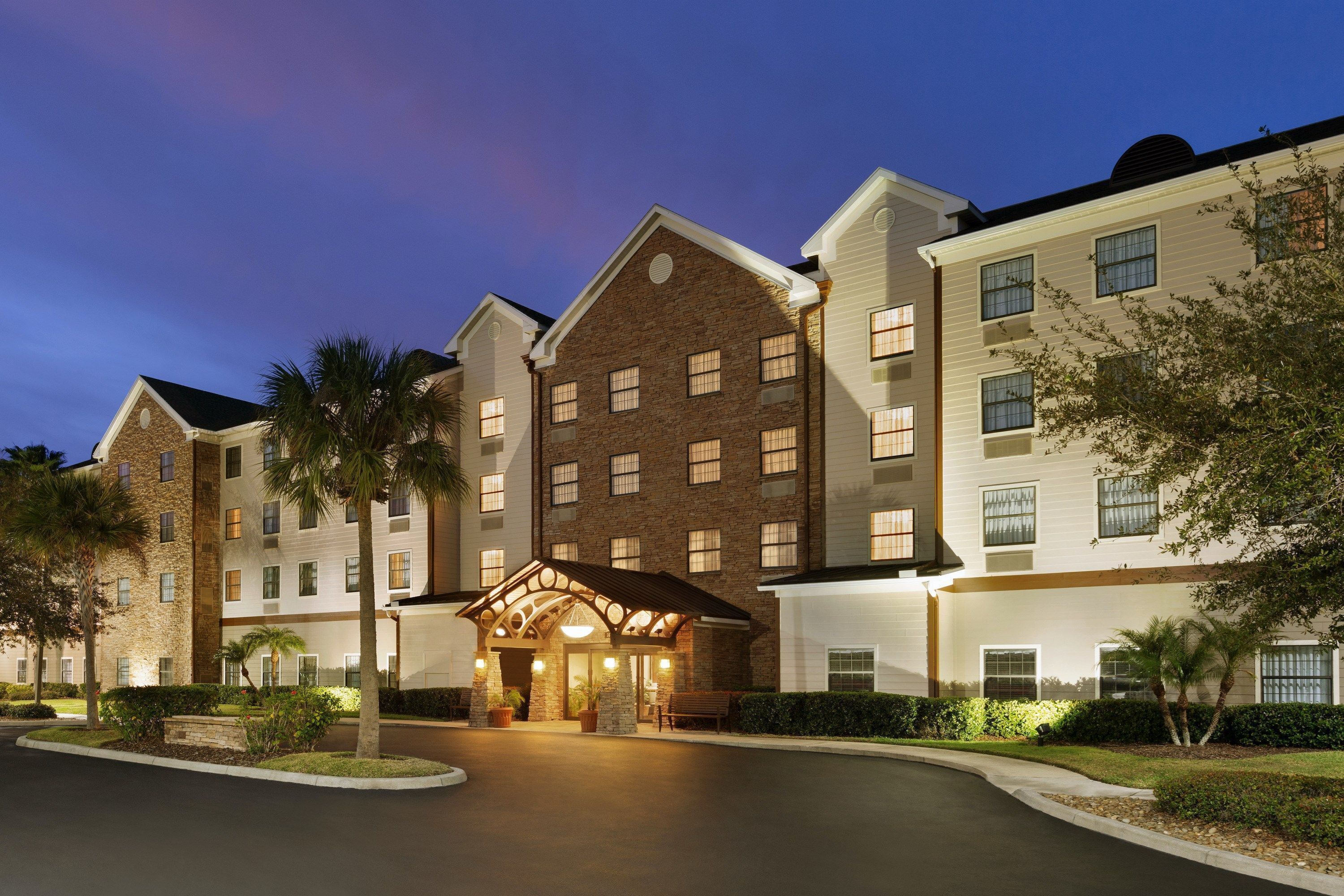 A multi-story hotel with a central entrance, surrounded by palm trees and a well-maintained lawn.