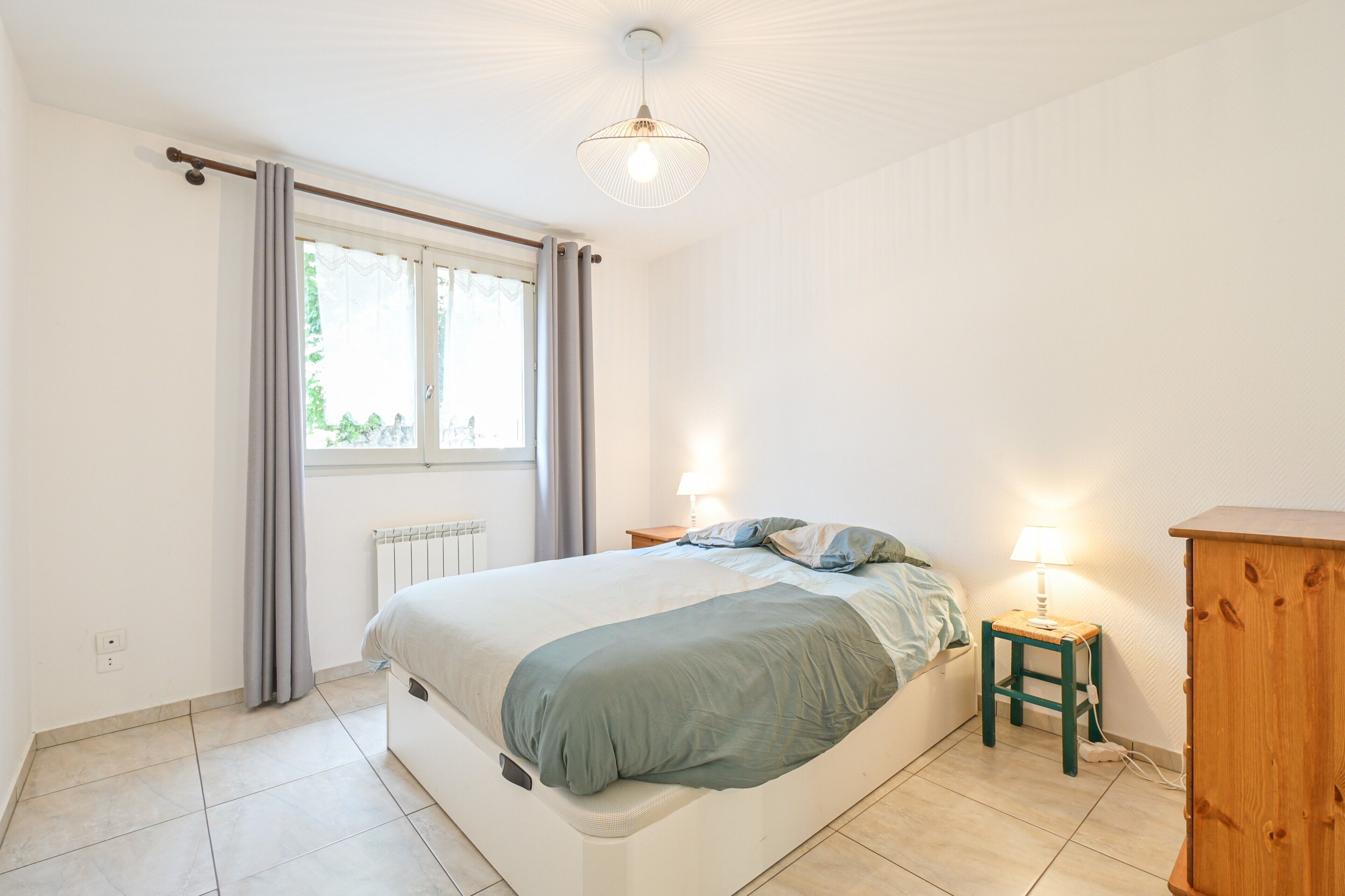 Une chambre à coucher avec un lit, des lampes de chevet, un radiateur, une fenêtre avec des rideaux et une armoire en bois.