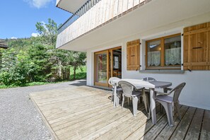Eine Holzterrasse mit Tisch und Stühlen, ein weißes Gebäude mit Holzhörnern und ein bewaldeter Hang im Hintergrund.