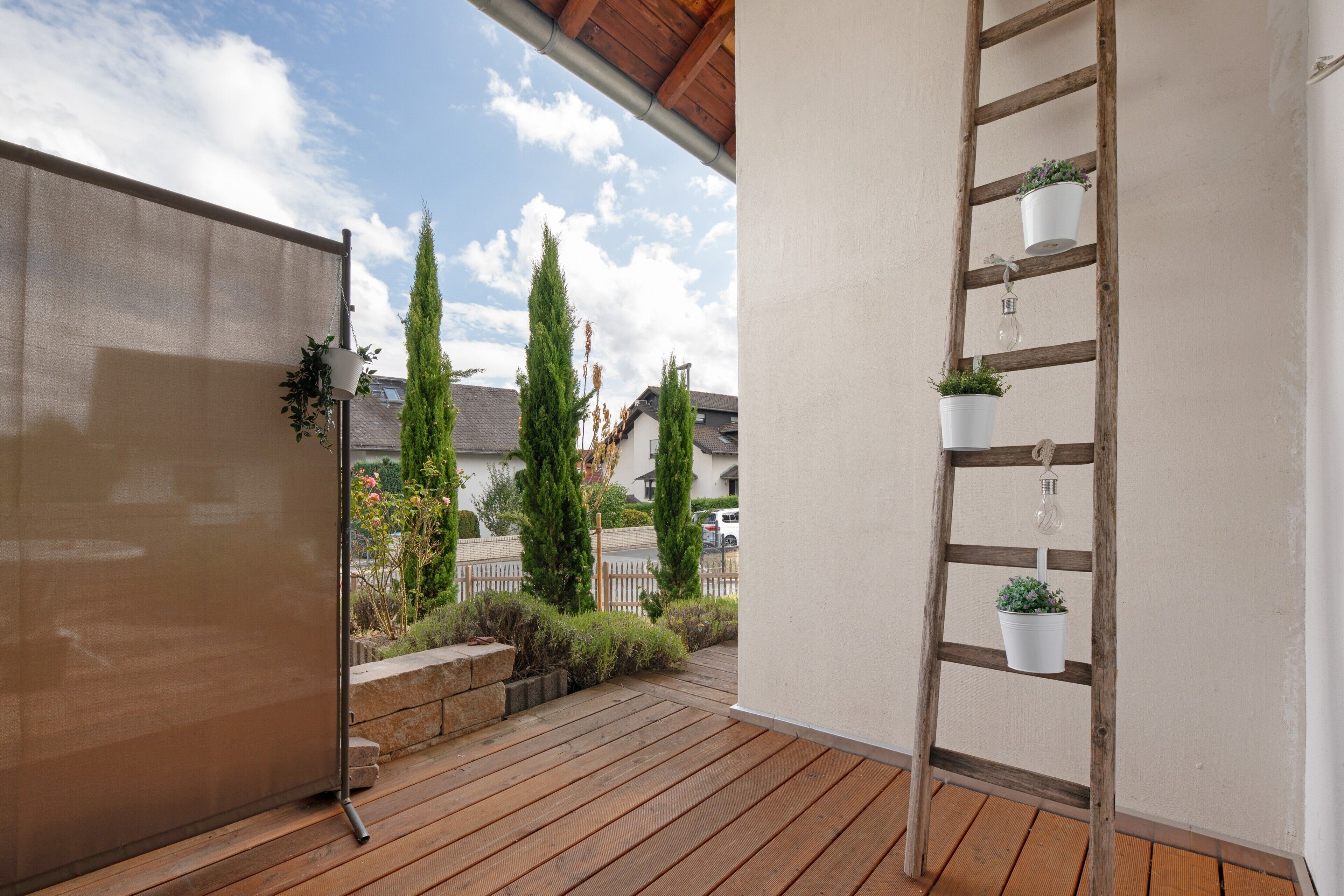 A wooden deck with a metal railing, potted plants, and a view of a residential area with houses and trees.