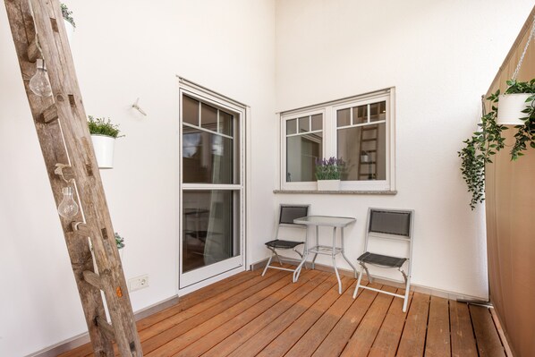 A wooden deck with a white wall, two windows, and a small table with two chairs.