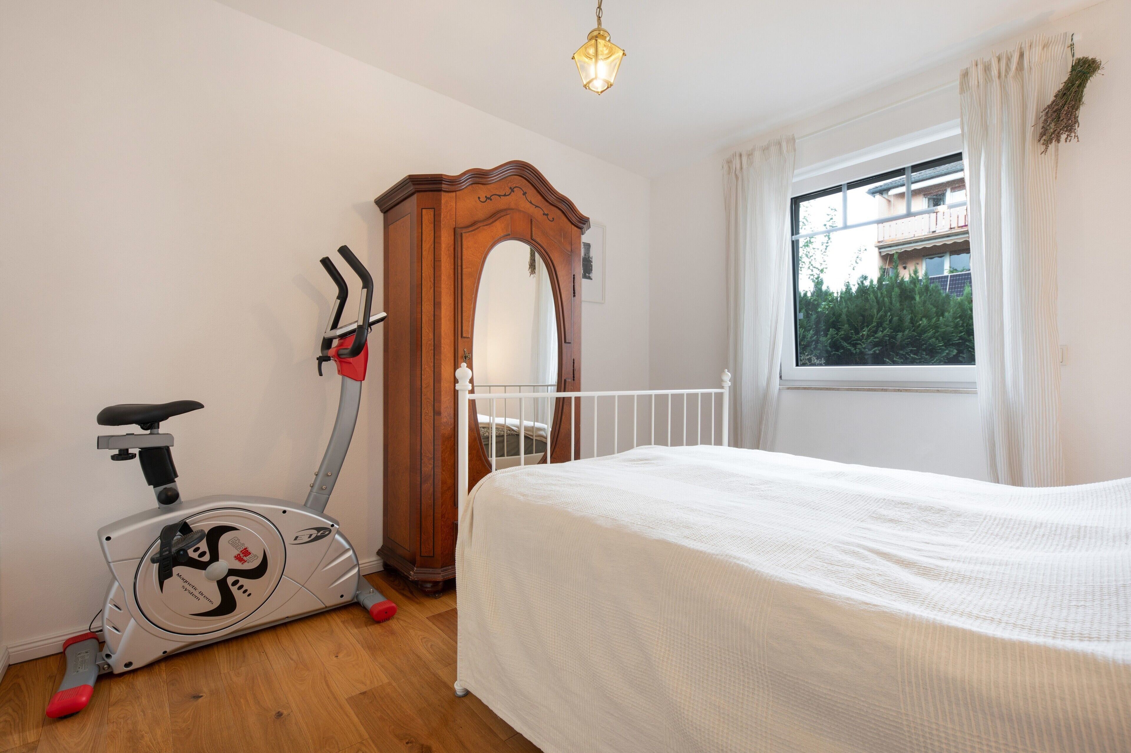 Un dormitorio con cama, bicicleta estática, armario de madera, ventana con vistas y lámpara colgante.