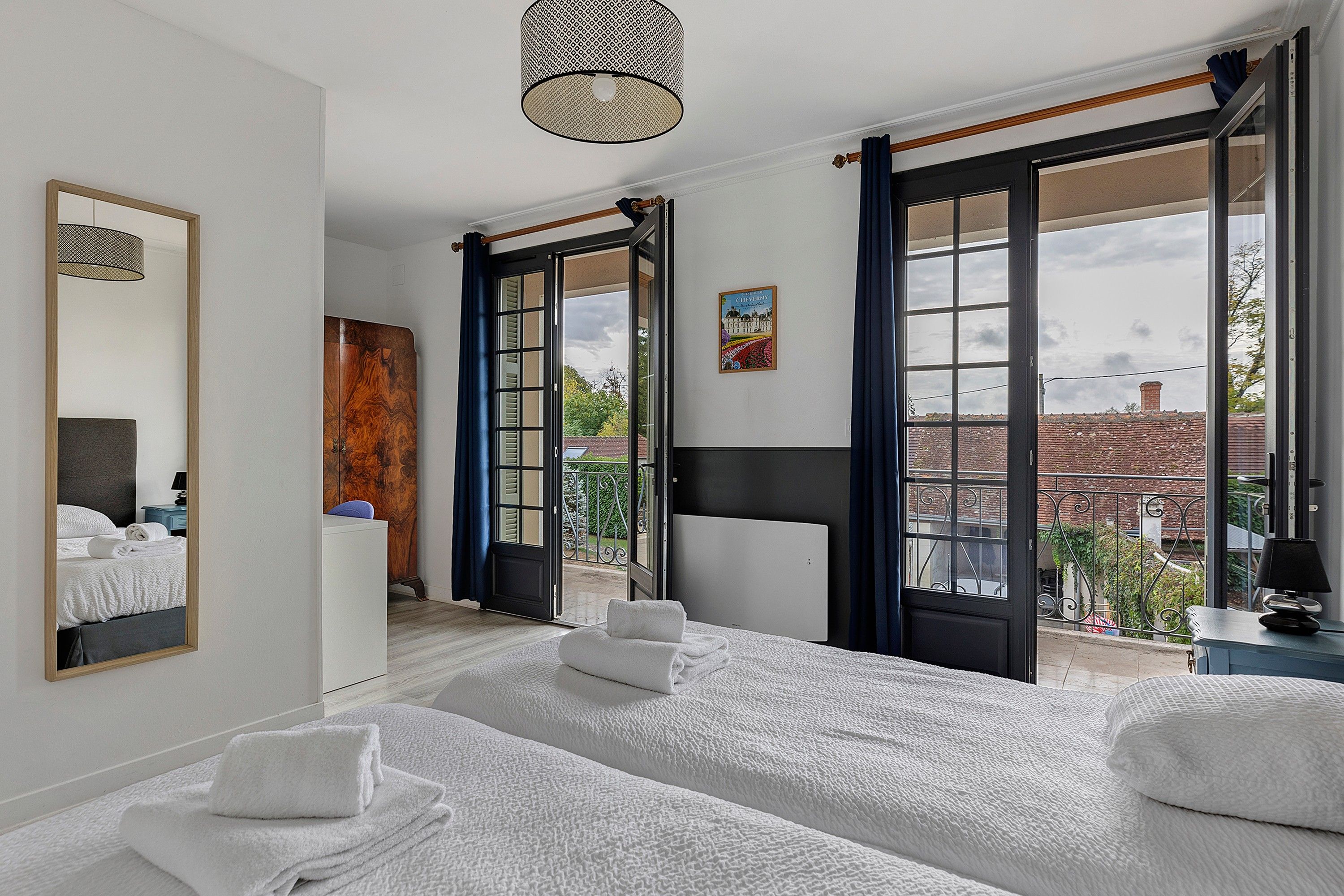 A hotel room with two beds, a large mirror, a wardrobe, and a balcony with a view of the city.
