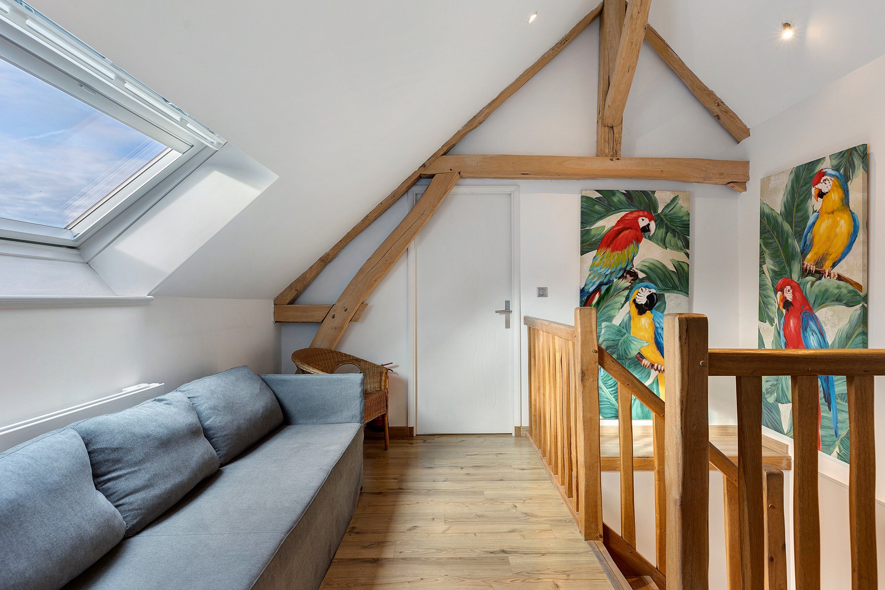 Une pièce avec un plafond en pente, un escalier en bois, un canapé et un tableau représentant des perroquets aux couleurs vives.