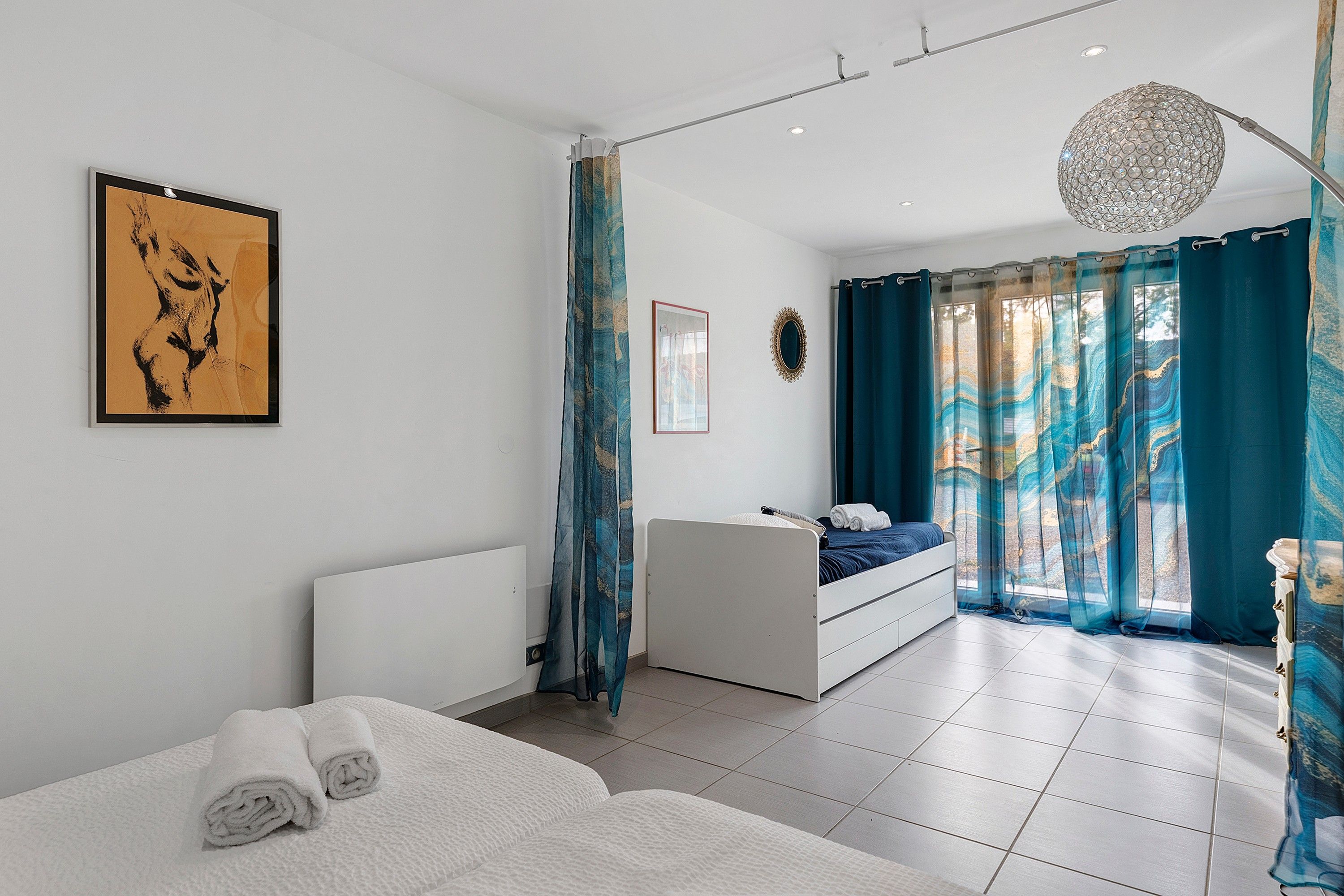 A modern bedroom with a bed, a bedside table, a dresser, a large window with curtains, and a ceiling light fixture.
