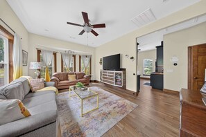 A spacious living room with a ceiling fan, a sofa, a coffee table, and a television on a cabinet. - Monte Ne Lakehouse - Lake View (Rogers)