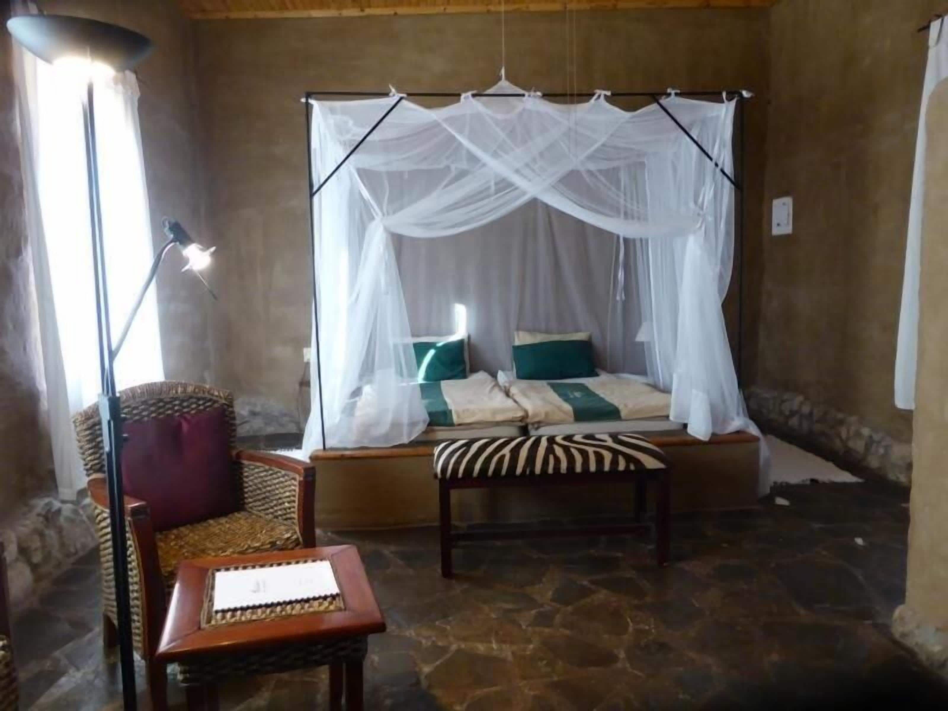 A room with a bed under a white canopy, a chair, and a table with a lamp.