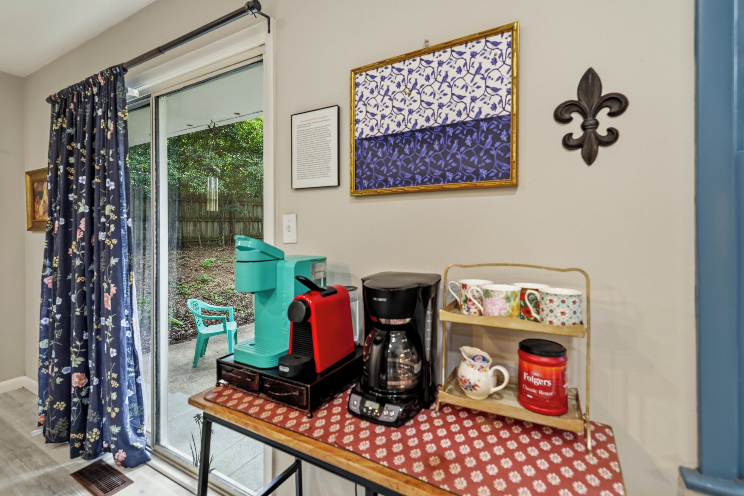 A room with a coffee station, a table with a red patterned tablecloth, a bookshelf with mugs, and a window with curtains.