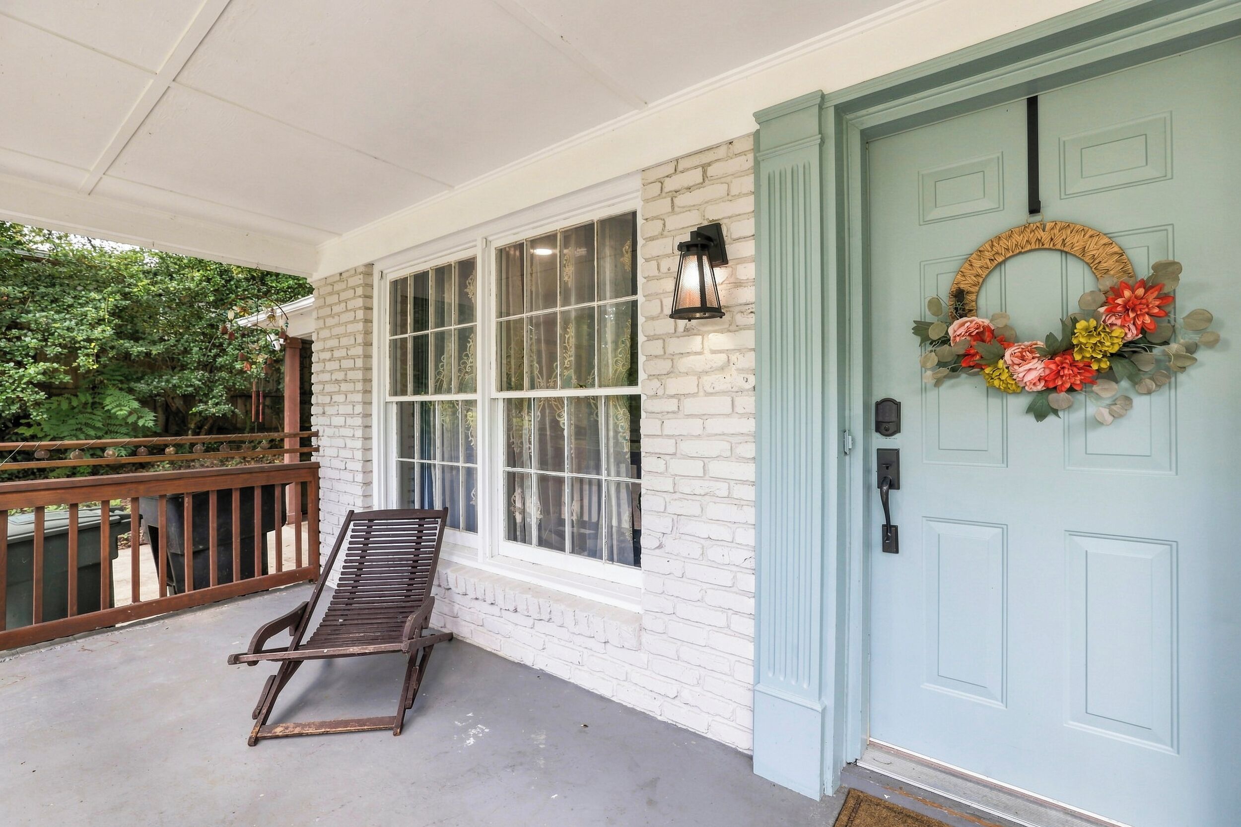 A porch with a wooden bench, a white brick wall, a blue door with a wreath, and a window with decorative curtains.