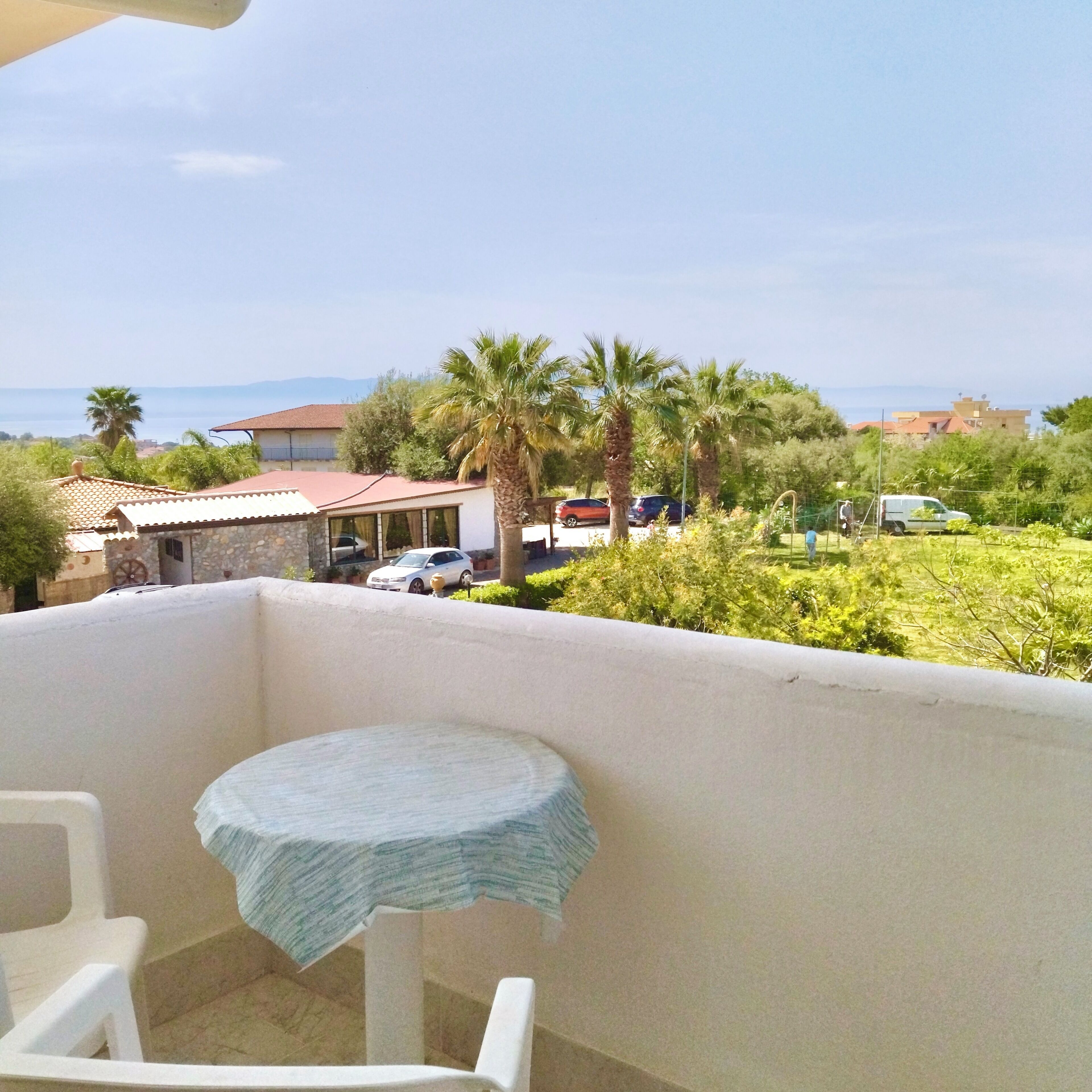 A balcony with a table and chairs, overlooking a residential area with parked cars and palm trees.