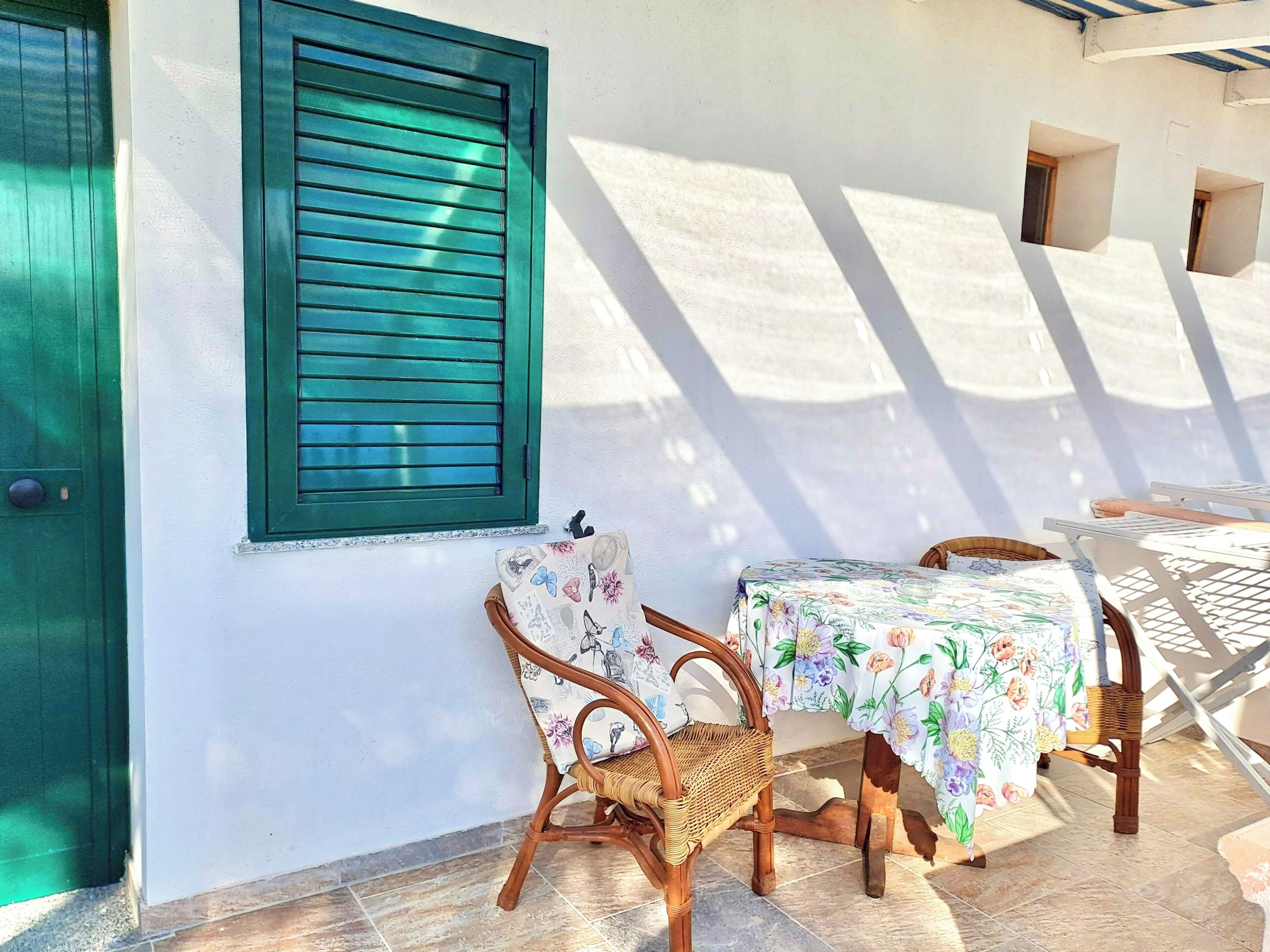 Une terrasse avec une table et des chaises, une porte verte et une fenêtre avec des volets.