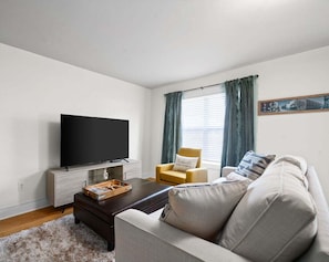 A living room with a flat-screen TV, a sofa, a coffee table, and a chair. - The Lofts of Downtown (540 N 2nd St, #210) (Clarksville)
