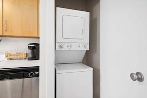 A compact kitchen with a dishwasher, a white washing machine, and wooden cabinets. - The Lofts of Downtown (540 N 2nd St, #210) (Clarksville)
