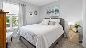 A bedroom with a bed, a bedside table, a window with curtains, a wall clock, and a painting. - The Lofts of Downtown (540 N 2nd St, #202) | Charming 2-Bedroom in the Heart of Clarksville, TN (Clarksville)