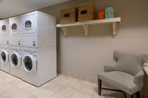 Lavandería con lavadoras y secadoras apiladas, una silla y estantes con cestas y recipientes.