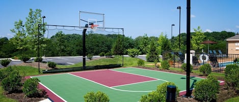 An outdoor basketball court with a green and red playing surface, surrounded by trees and a fence.