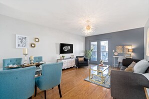 Uma sala de estar moderna com área de jantar, apresentando uma TV de tela plana, poltronas estofadas em azul, um sofá cinza e piso de madeira.
