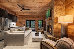 A modern interior with a stone fireplace, a sofa set, a dining area, and a kitchen with a stainless steel refrigerator. - Hemlock Haven: Creekside Paradise • Pool • Sauna • HotTub • FirePit • Fun Games (Blue Ridge)